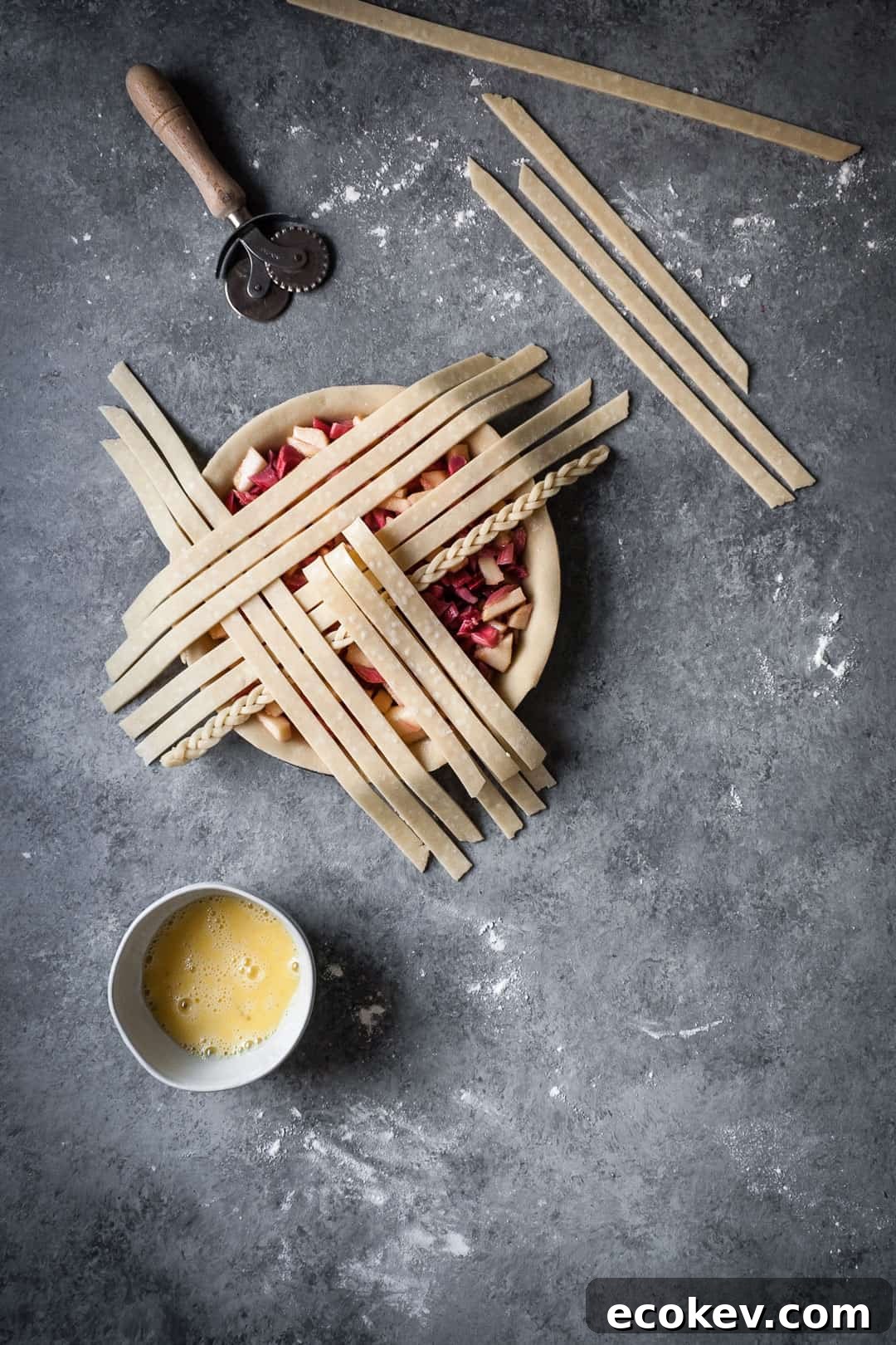 process of making a rhubarb apple pie with ginger and lemongrass - pie crust with apple rhubarb filling and lattice top in process with egg wash and dough cutter nearby