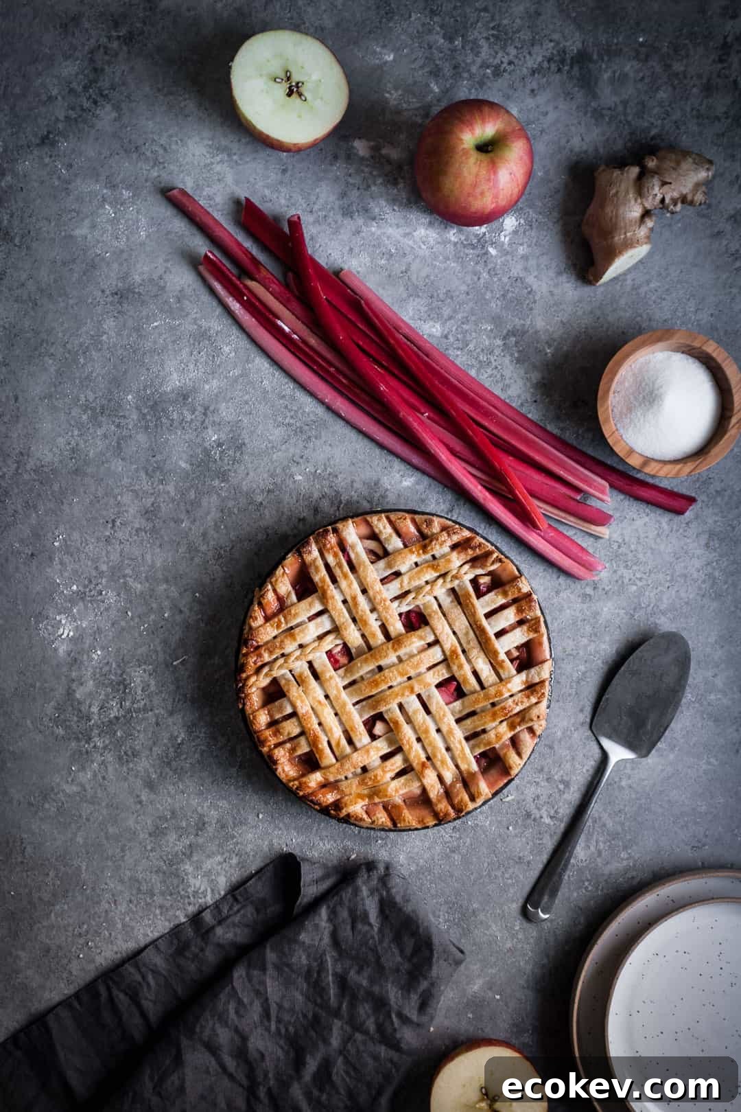 grey surface with wide view of pie making ingredients for rhubarb apple pie with ginger and lemongrass including apples rhubarb and ginger along with pie server and empty white plates