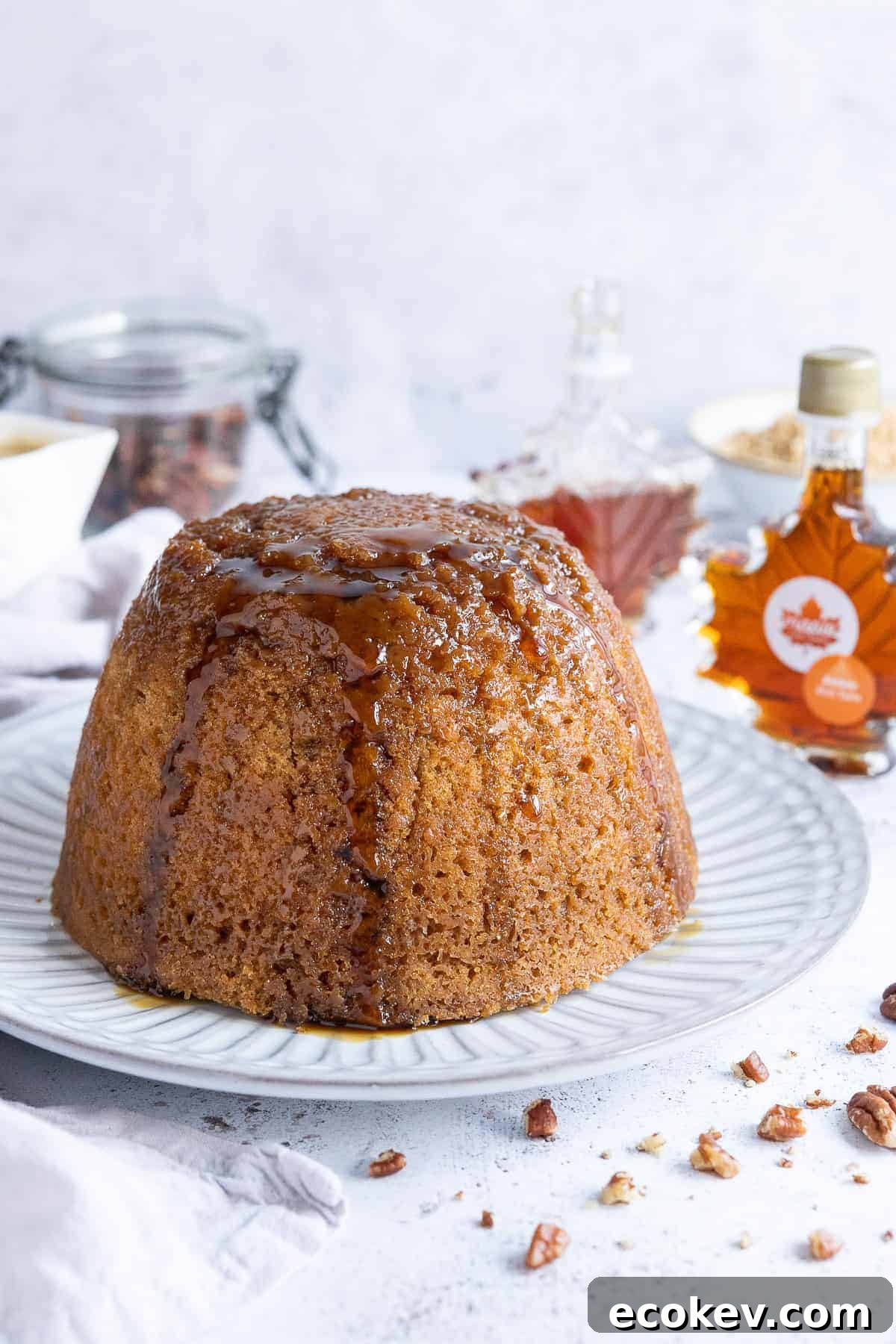 Vegan steamed maple syrup pudding on a white plate with bottles of maple syrup, a jug of custard and a jar on pecans in the background.