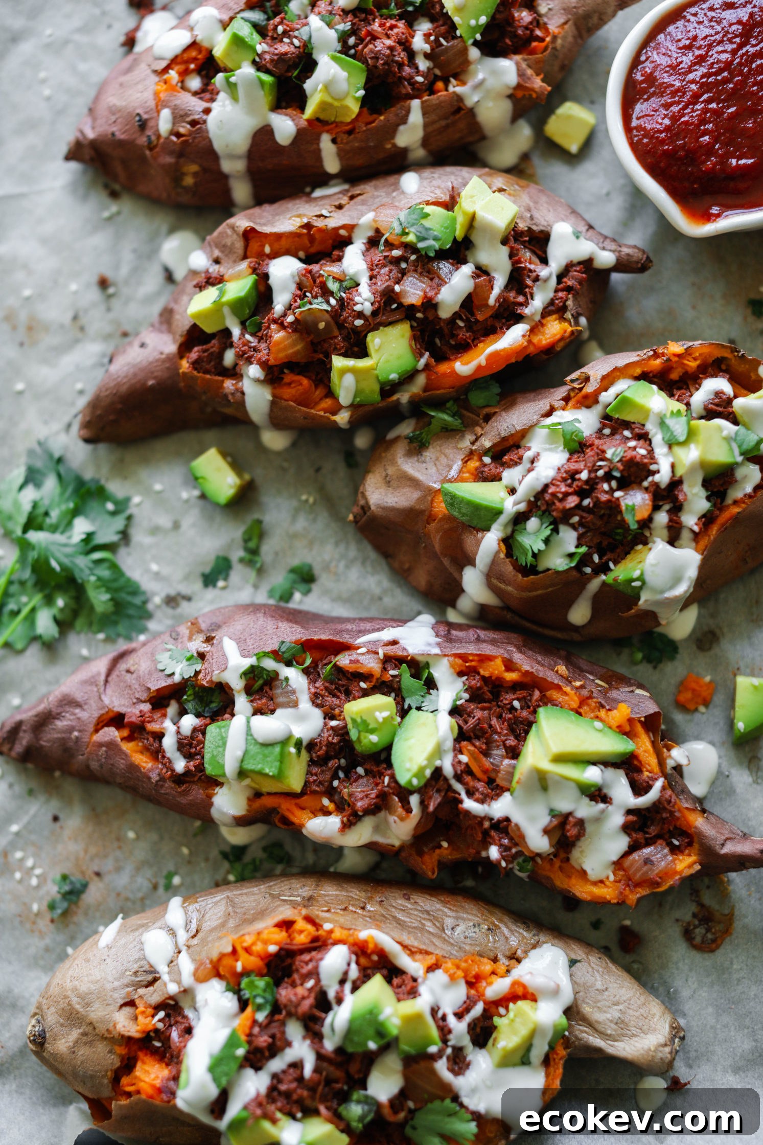 BBQ Jackfruit Stuffed Sweet Potatoes on parchment paper topped with yogurt tahini, cilantro, and avocado with BBQ sauce on side