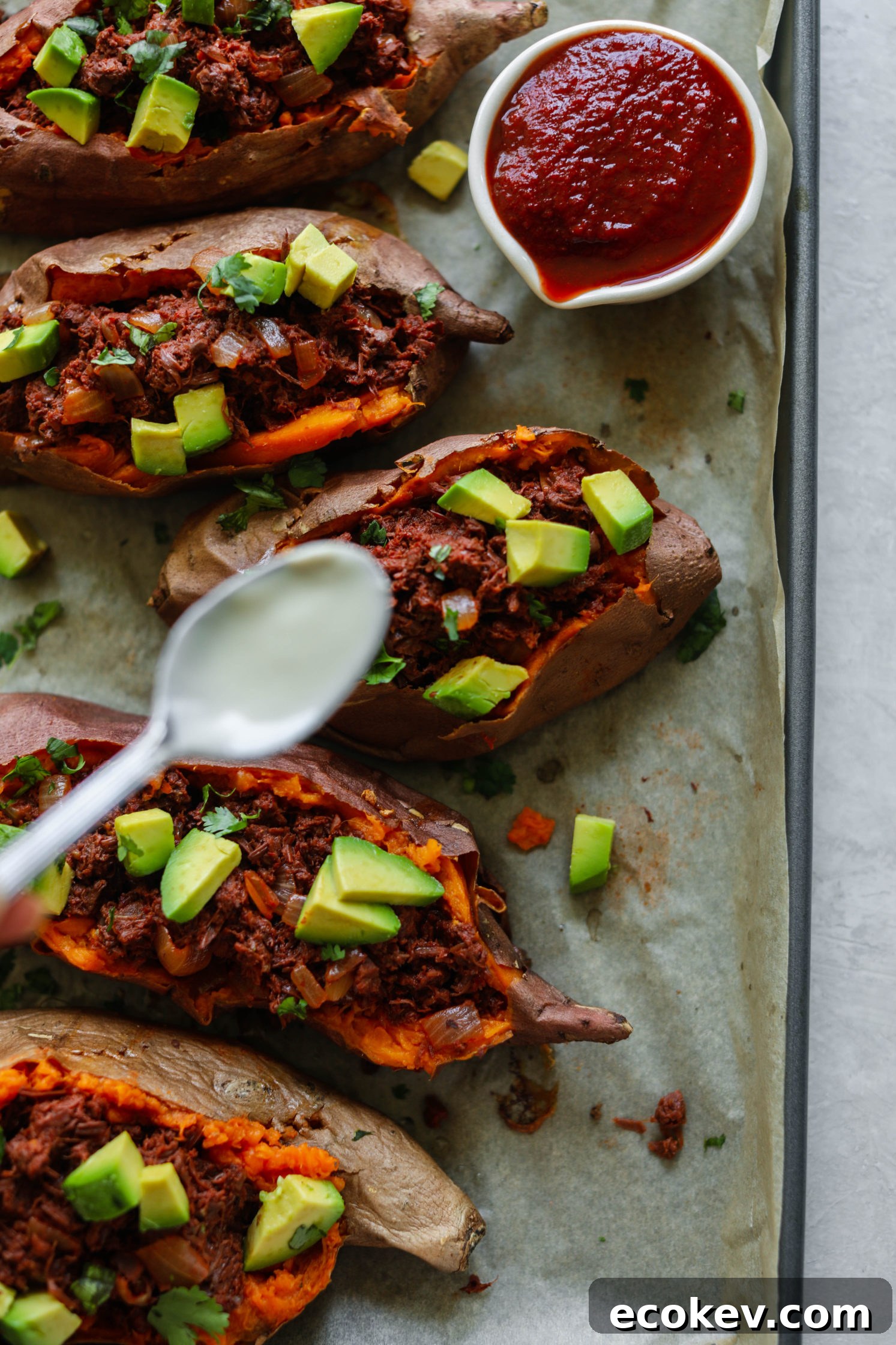 BBQ Jackfruit Stuffed Sweet Potatoes on parchment paper topped with spoon of yogurt tahini overhead and bowl of BBQ sauce on side