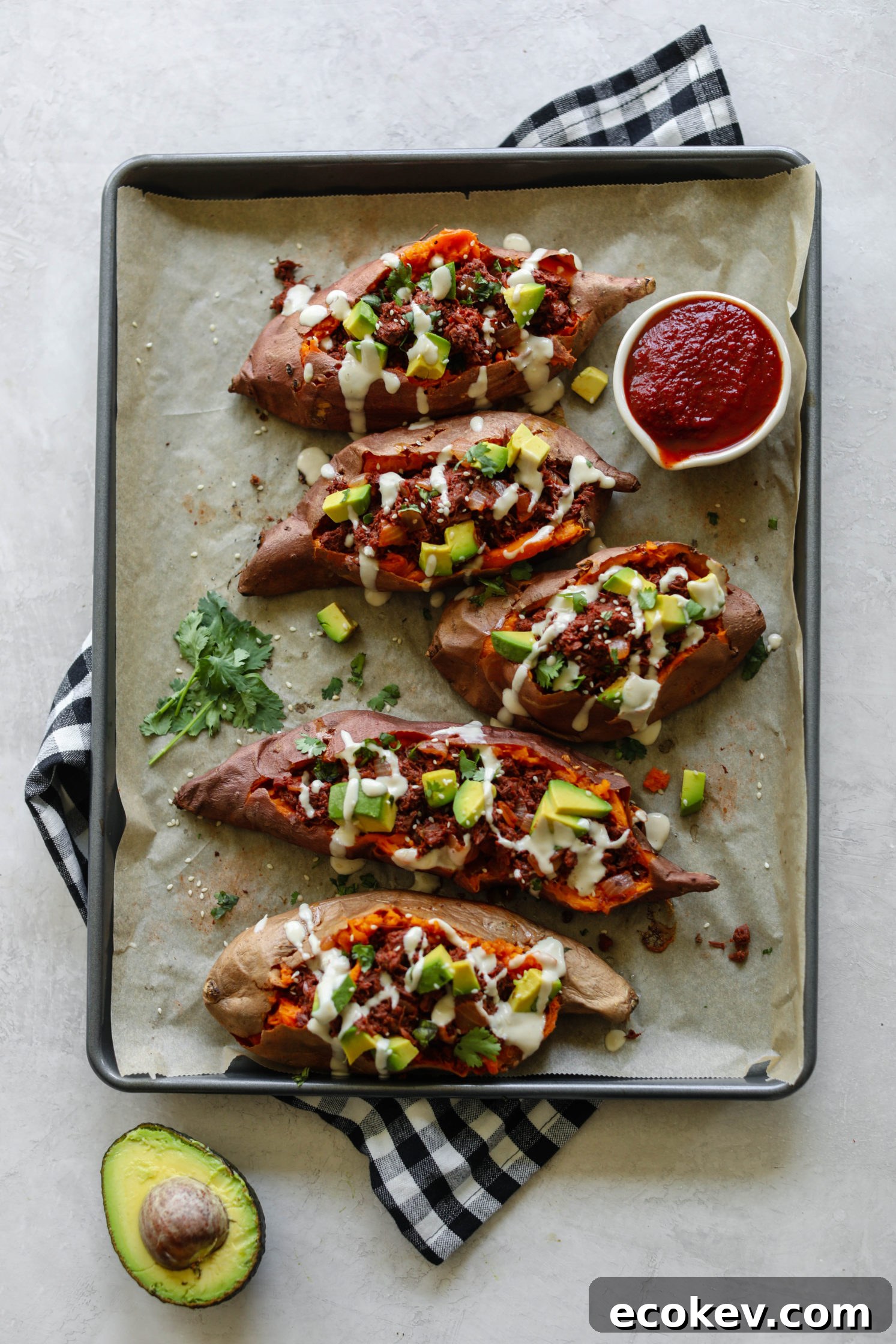 BBQ Jackfruit Stuffed Sweet Potatoes on parchment paper topped with yogurt tahini, cilantro, and avocado with BBQ sauce on side