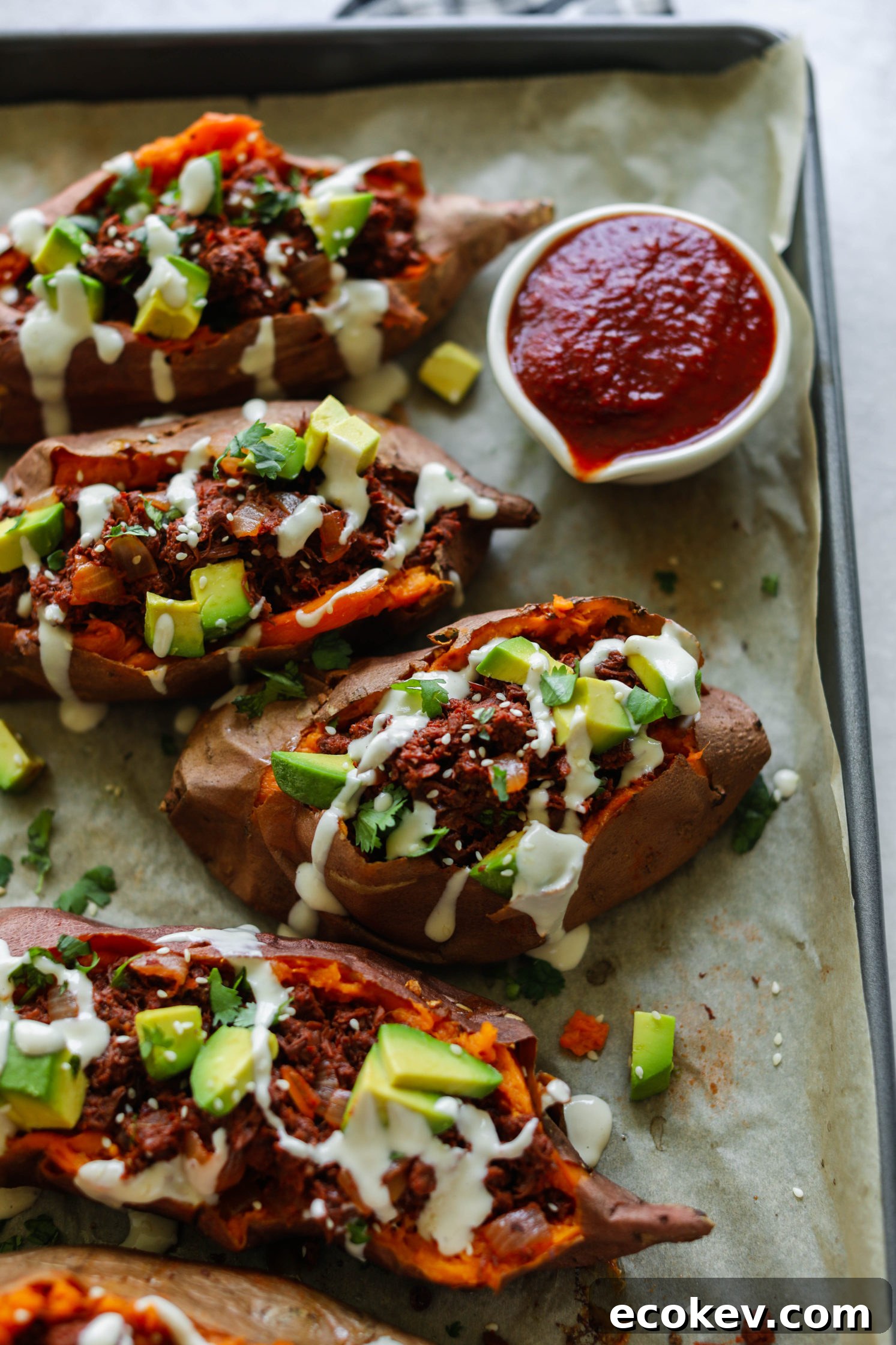 BBQ Jackfruit Stuffed Sweet Potatoes on parchment paper topped with yogurt tahini, cilantro, and avocado with BBQ sauce on side