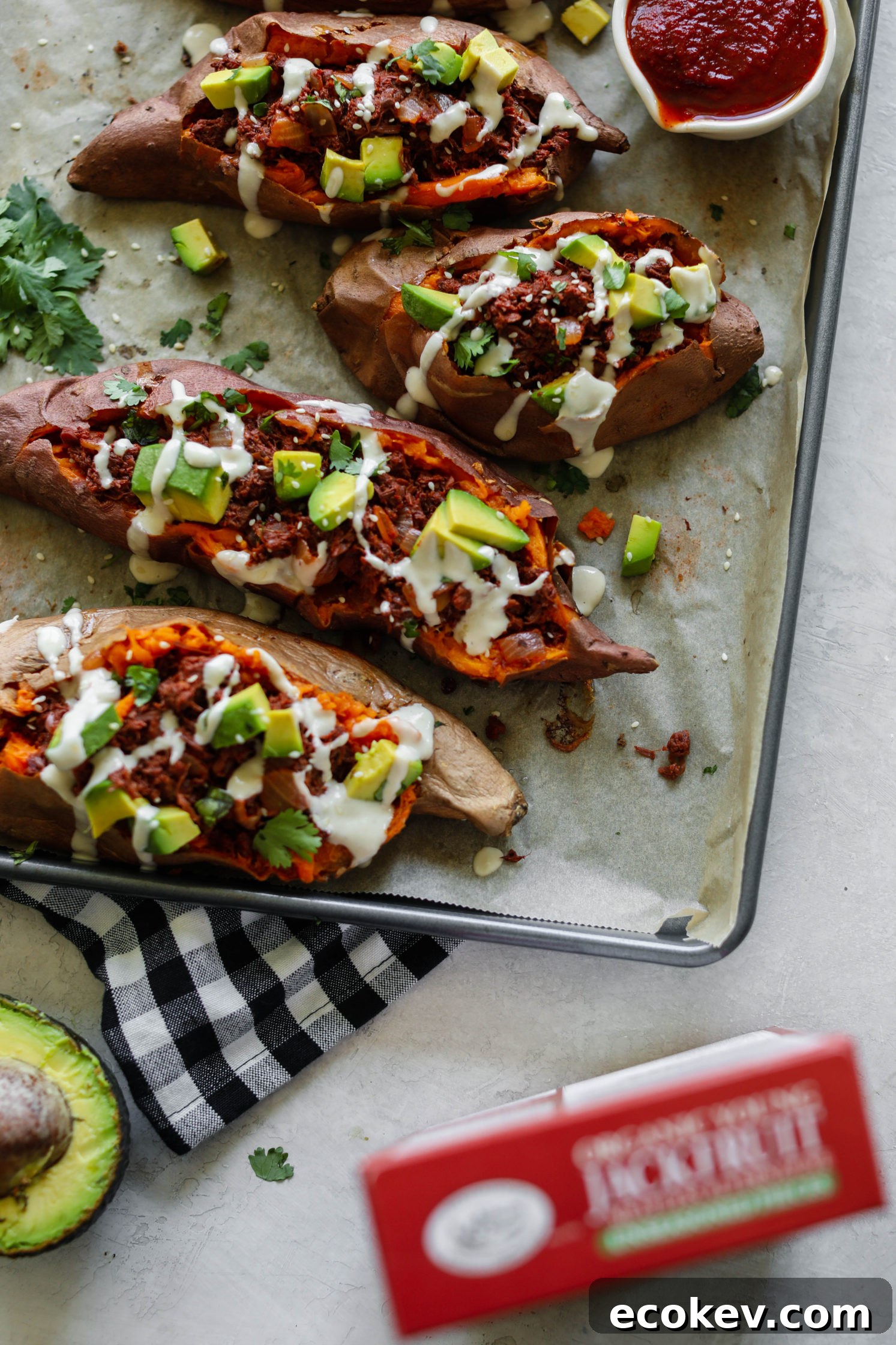 BBQ Jackfruit Stuffed Sweet Potatoes on parchment paper topped with yogurt tahini, cilantro, and avocado with BBQ sauce on side
