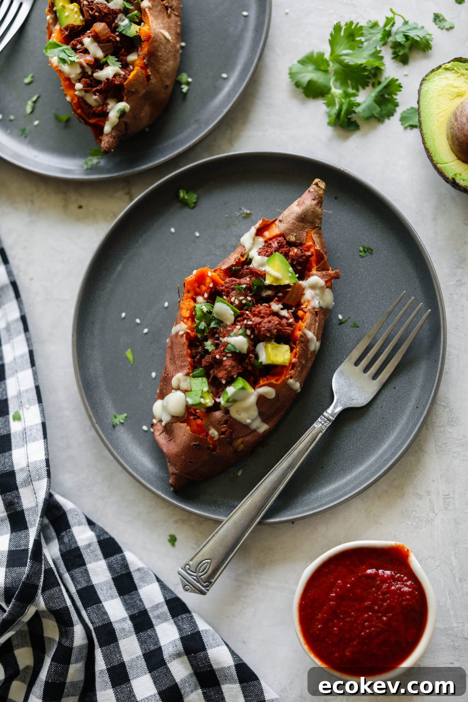 BBQ Jackfruit Stuffed Sweet Potatoes on plate topped with yogurt tahini, cilantro, and avocado with BBQ sauce on side