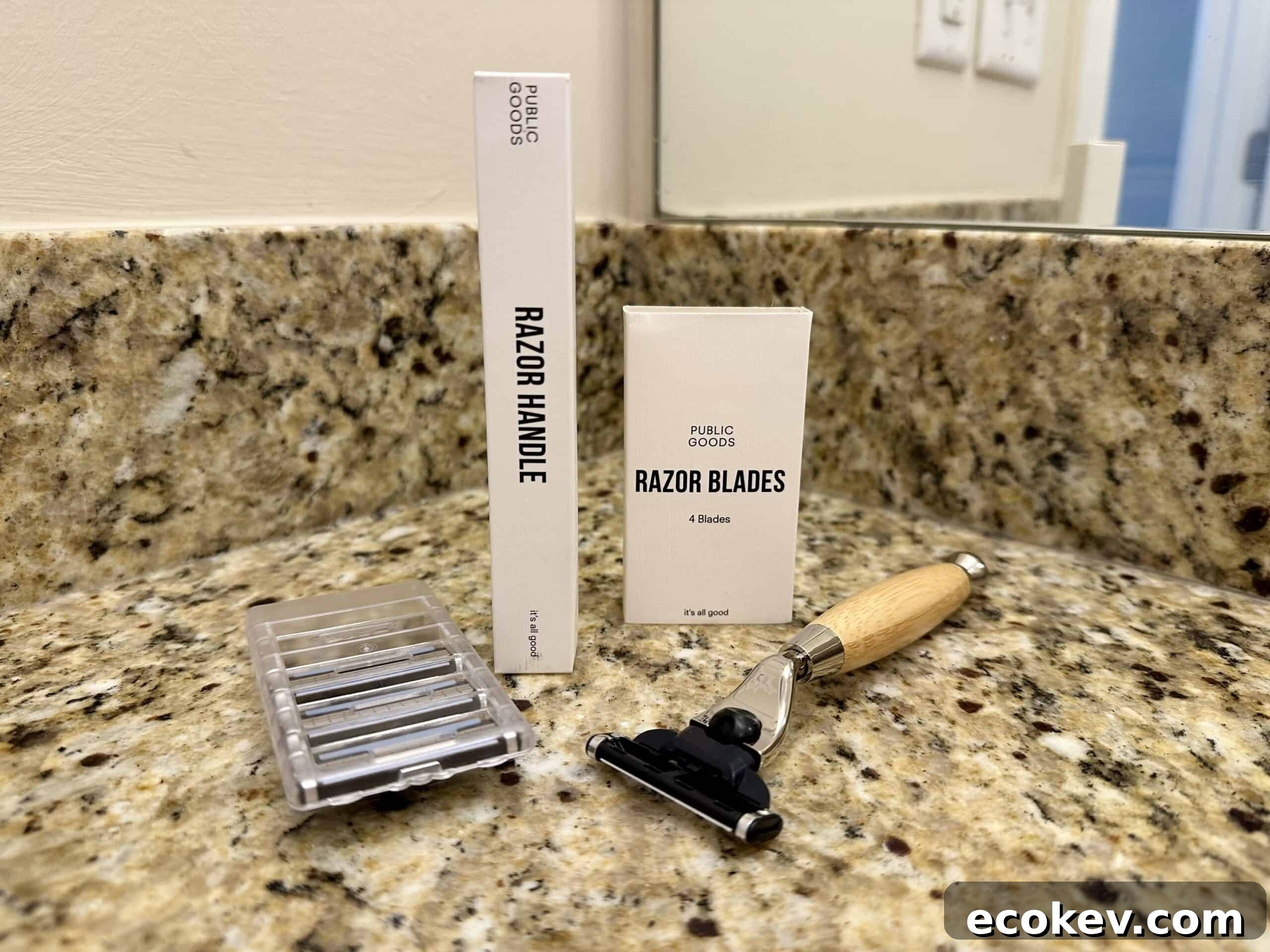 A Public Goods razor set displayed on a granite bathroom countertop, including a wooden-handled razor, a box labeled “Razor Handle,” a box labeled “Razor Blades,” and a plastic case holding replacement blades. A mirror and wall outlet are visible in the background.