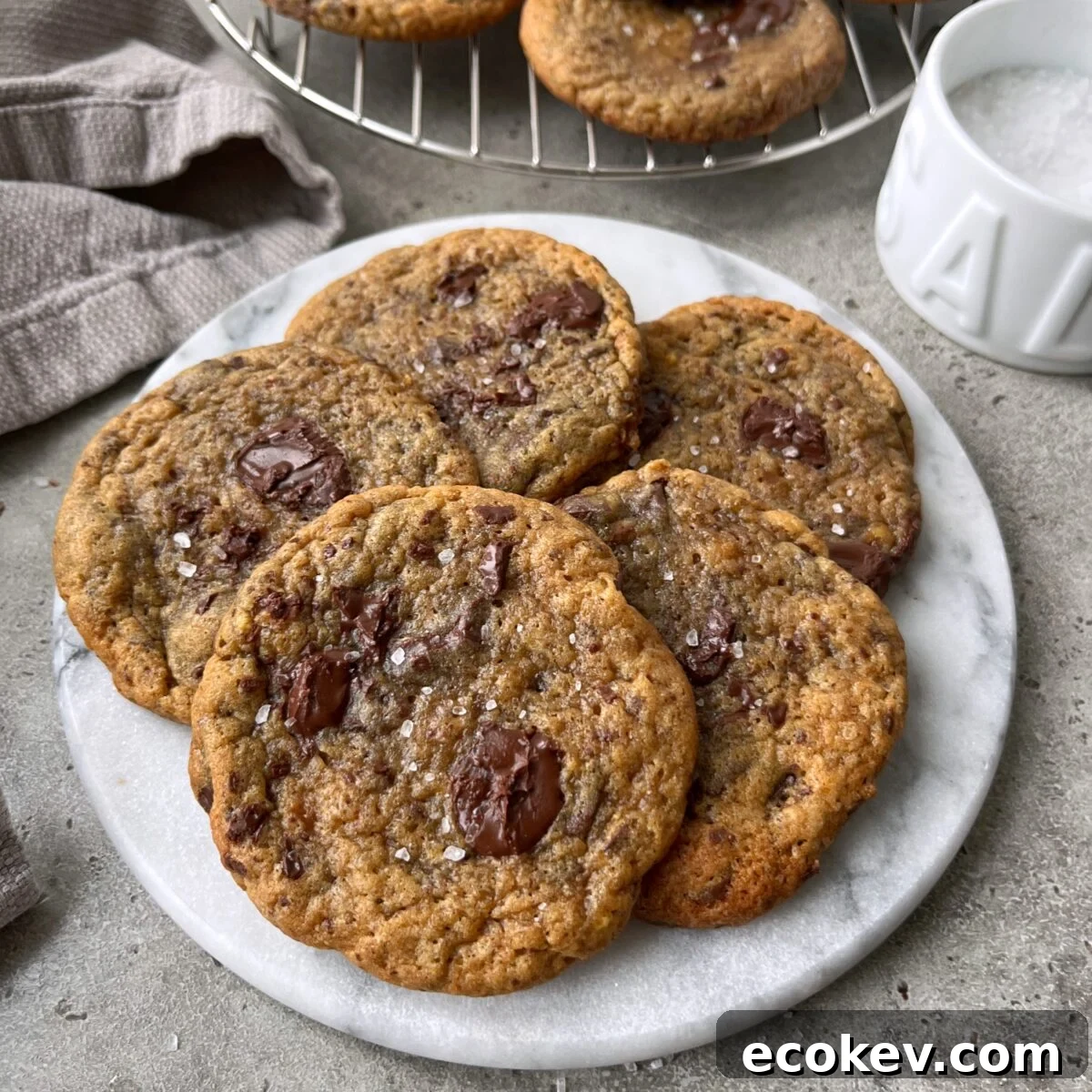 A plate of banana bread cookies topped with sea salt with a cooling rack of cookies in the background.