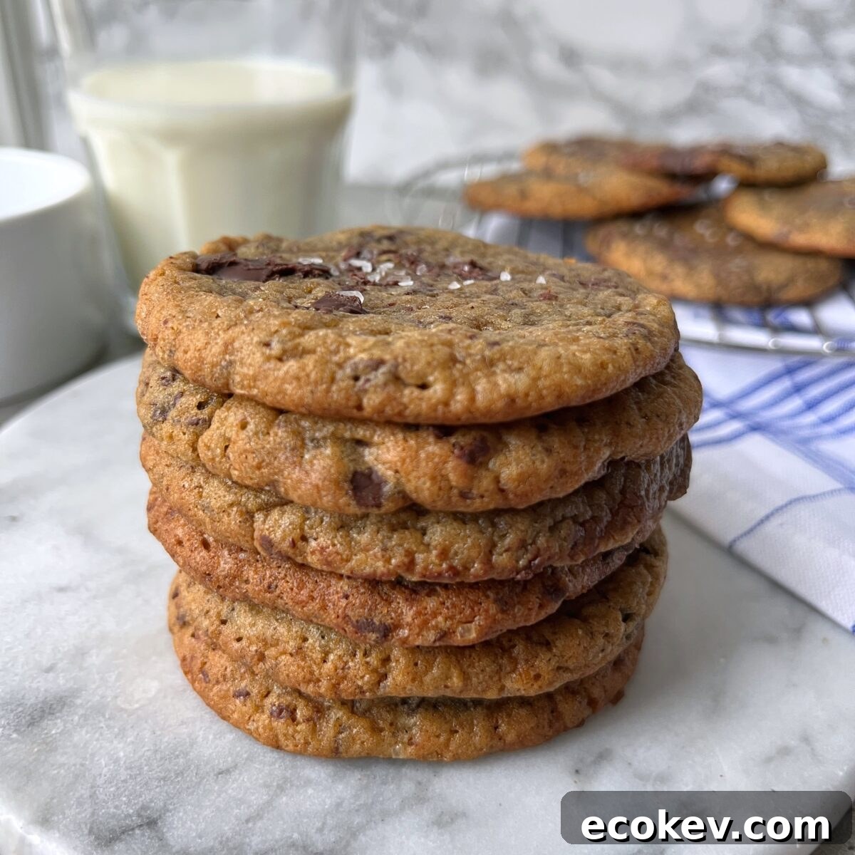 A stack of banana bread cookies sits on a marble surface, with a glass of milk and more cookies in the background.