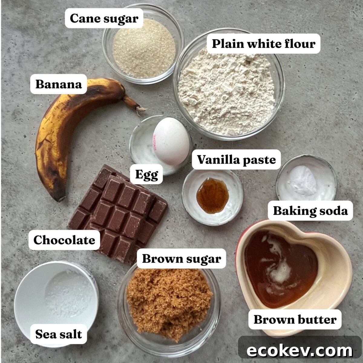 Banana bread cookie ingredients on a gray surface, including a banana, chocolate, egg, vanilla paste, brown and cane sugar, sea salt, plain flour, baking soda, and brown butter.