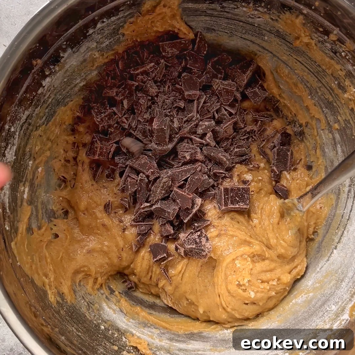 A metal bowl filled with cookie dough topped with chunks of chopped dark chocolate, being mixed with a metal spoon.