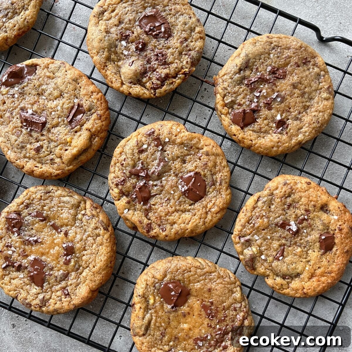 Eight chocolate chip cookies with visible chocolate chunks and sprinkled sea salt cooling on a black wire rack.