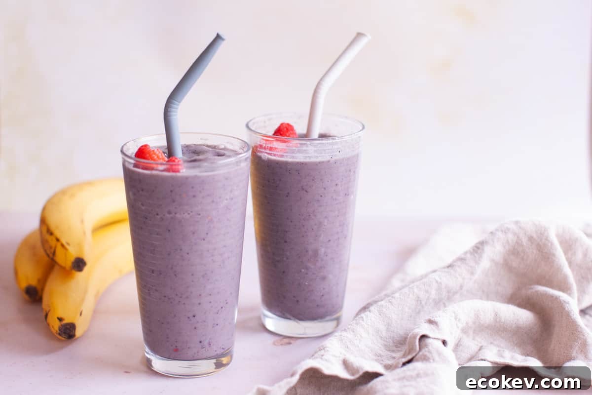 A wide shot of two glasses with straws filled with the smoothie topped with fresh raspberries next to two bananas.