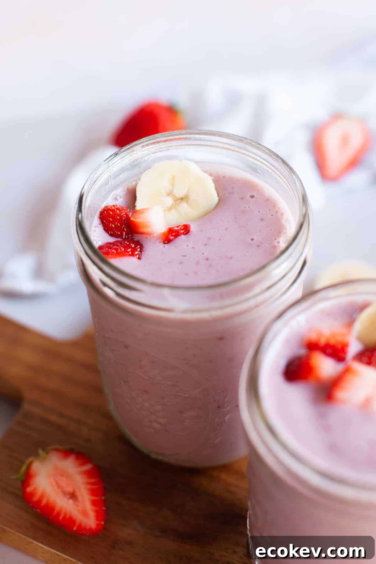 Two jars filled with pink fruit smoothie topped with banana slices and strawberry pieces, placed on a wooden surface with additional strawberries in the background.